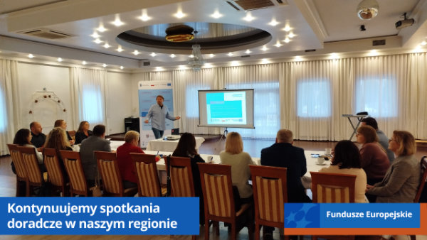 Sala konferencyjna. Uczestnicy spotkania siedzą wzdłuż dużego stołu. Na przeciwko prowadzący i ekran rzutnika