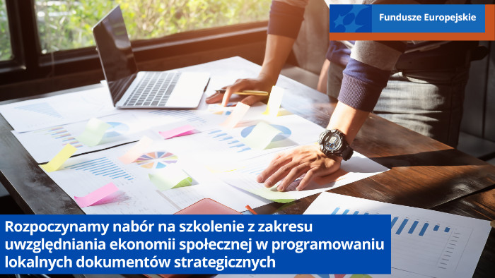 Mężczyzna stoi przy biurku. Na biurku leży laptop i są rozłożone różne dokumenty